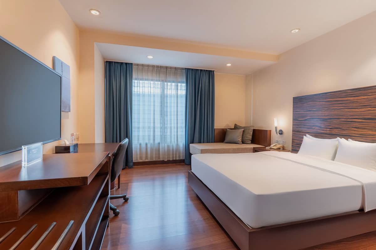 Habitación de hotel con una cama grande, un escritorio, televisión y ventana con cortinas.