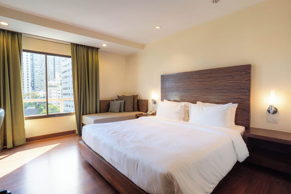 Habitación de hotel con una cama grande, un sofá y un escritorio. Cuenta con una ventana con cortinas, una lámpara de pared y vistas a edificios.