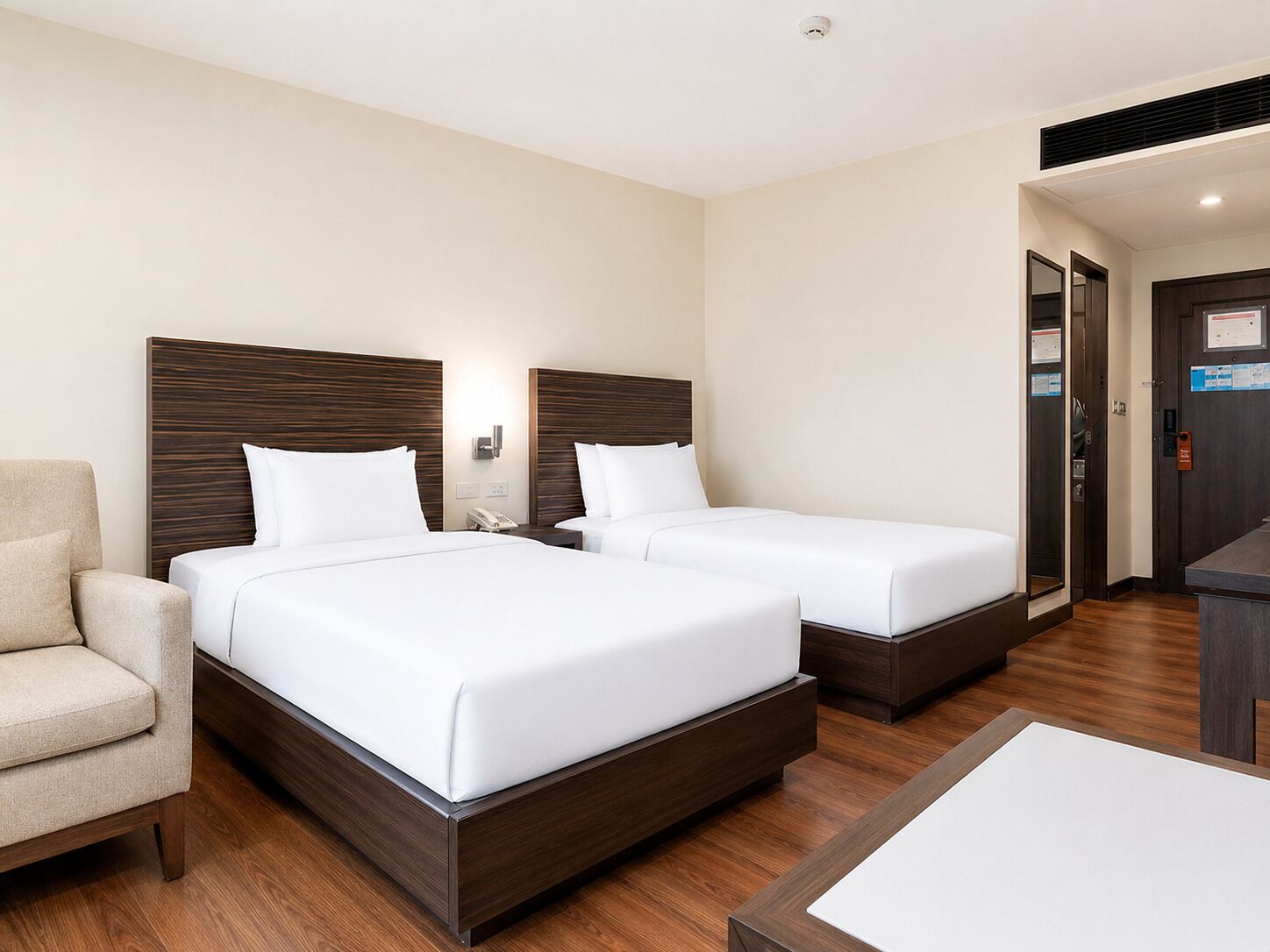 A hotel room with two beds, a chair, a desk, and a door.