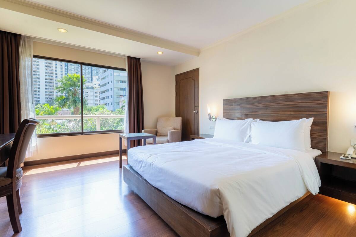 Habitación de hotel con una cama grande, un escritorio, una silla, una mesita de noche y una ventana con vistas a edificios y vegetación.