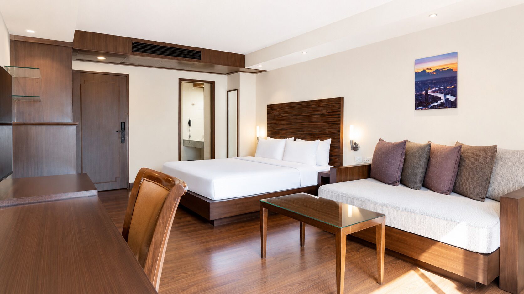 A hotel room with a bed, a sofa, a dining table, and a view of a cityscape.