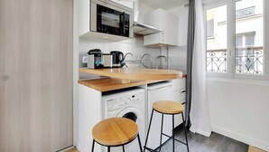 Ett kompakt kök med mikrovågsugn, kaffebryggare och tvättmaskin, trästolar och utsikt över balkongen.