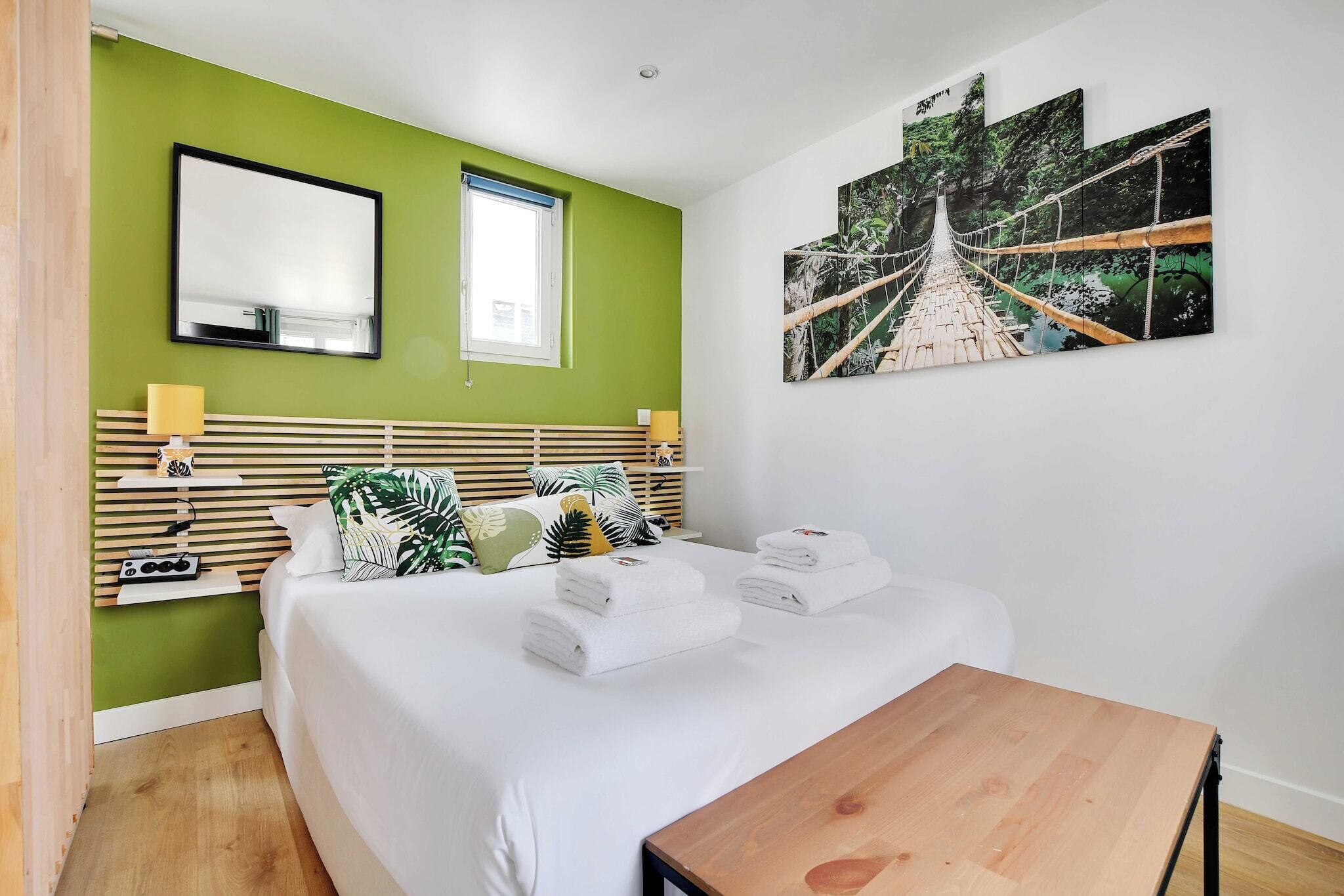 Habitación de hotel con cama, cabecera de madera, mesita de noche, espejo, ventana y un cuadro grande con un puente.