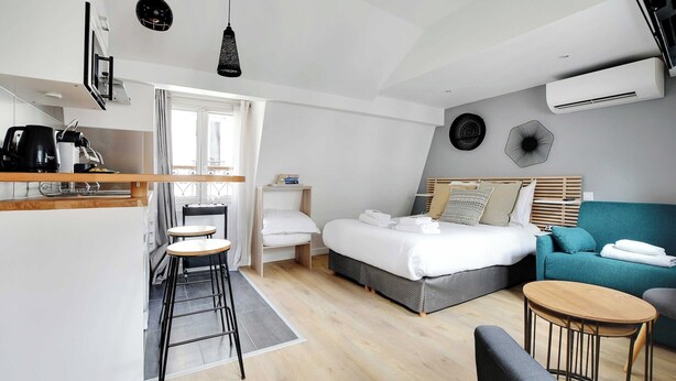 A modern hotel room with a bed, a sofa, a small table, a kitchen area, and an air conditioning unit. - Quiet studio - 2P - Sentier (Paris)