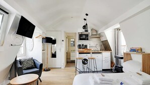 A modern hotel room with a bed, a desk, a chair, a TV mounted on the wall, a kitchen area, and a view of the hallway. - Quiet studio - 2P - Sentier (Paris)