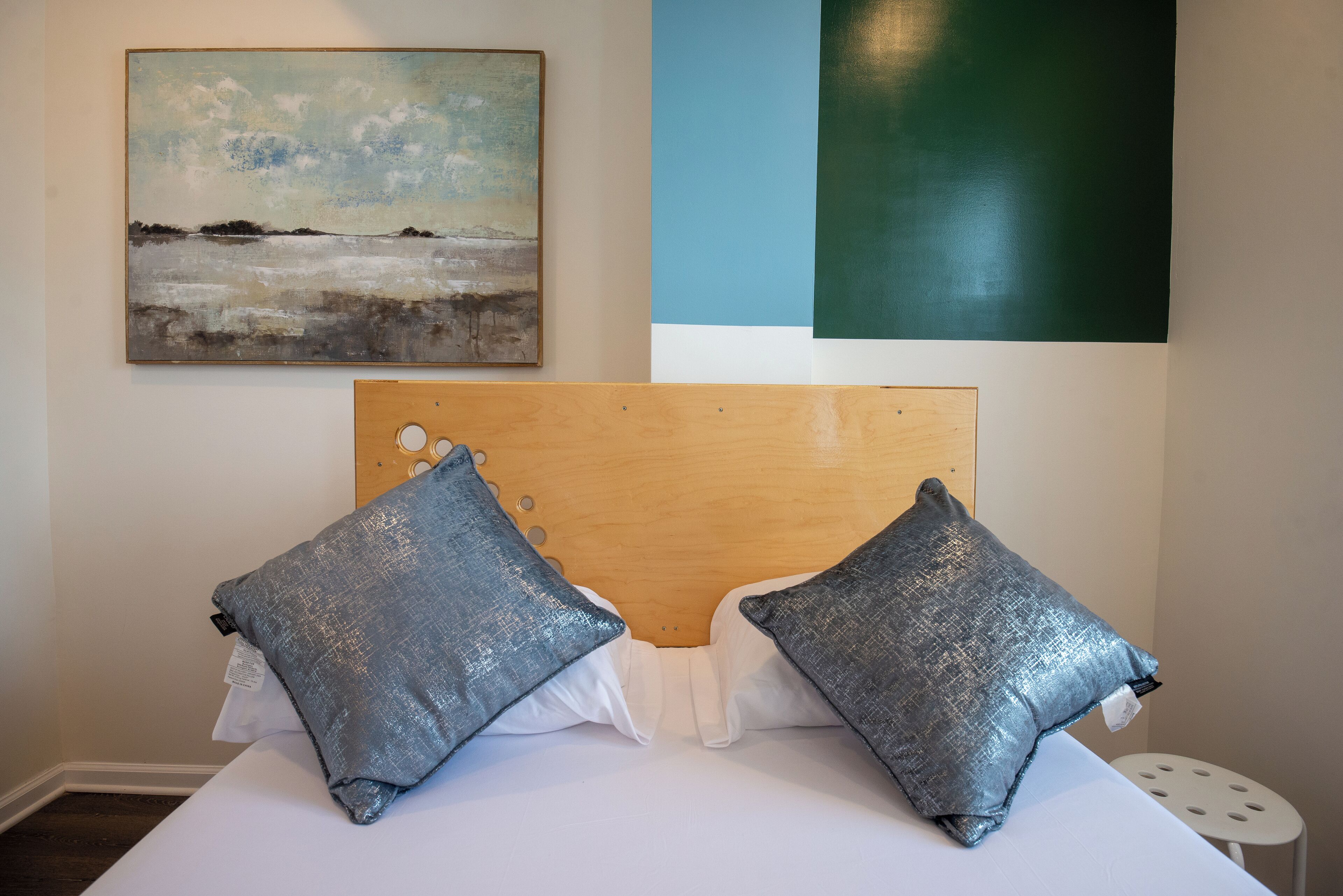 A bed with a wooden headboard, two grey pillows, and a white pillow. There is a painting on the wall above the bed.