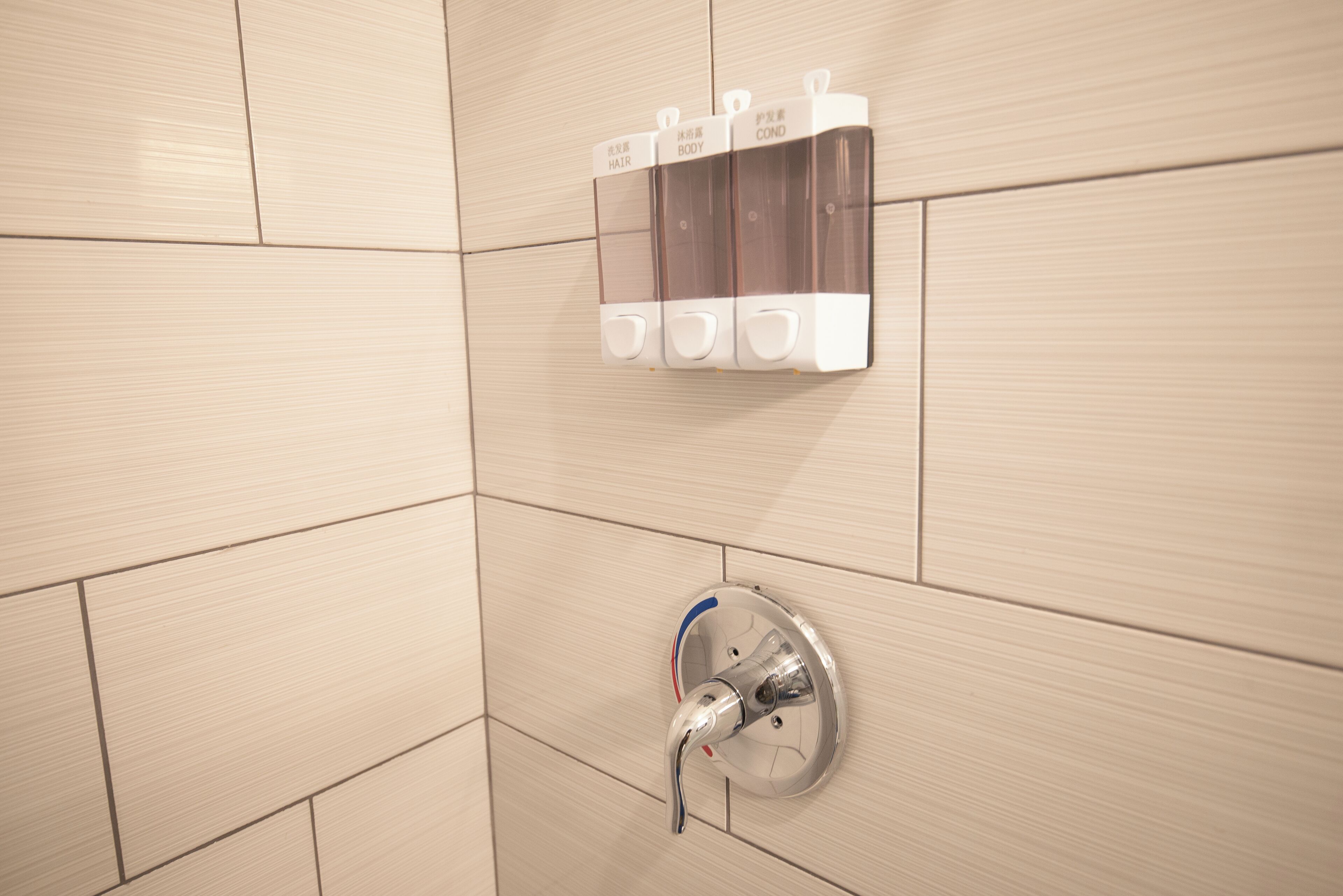 A bathroom with beige tiles, a wall-mounted soap dispenser, and a chrome faucet.