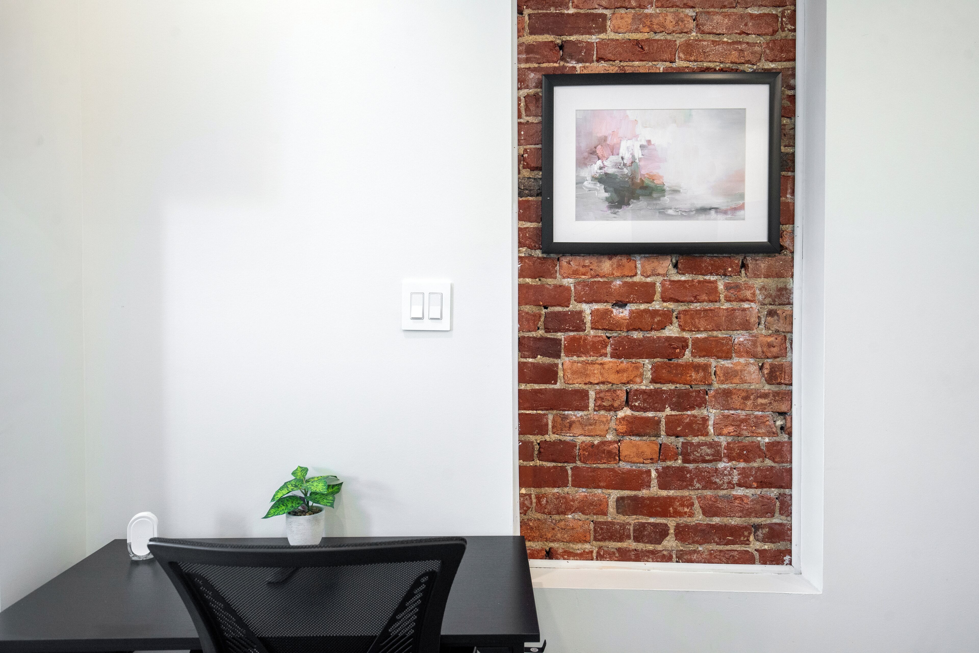 A room with a brick wall, a framed picture, a small table with a chair, and a potted plant.