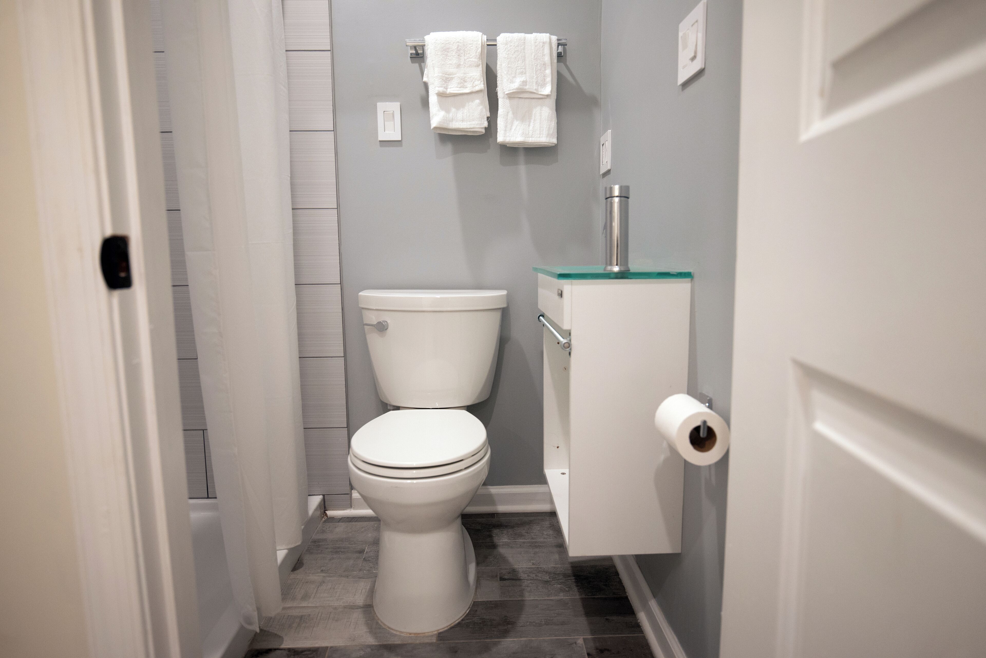 A small, modern bathroom with a toilet, a glass-topped sink, and two white towels hanging on a towel rack.