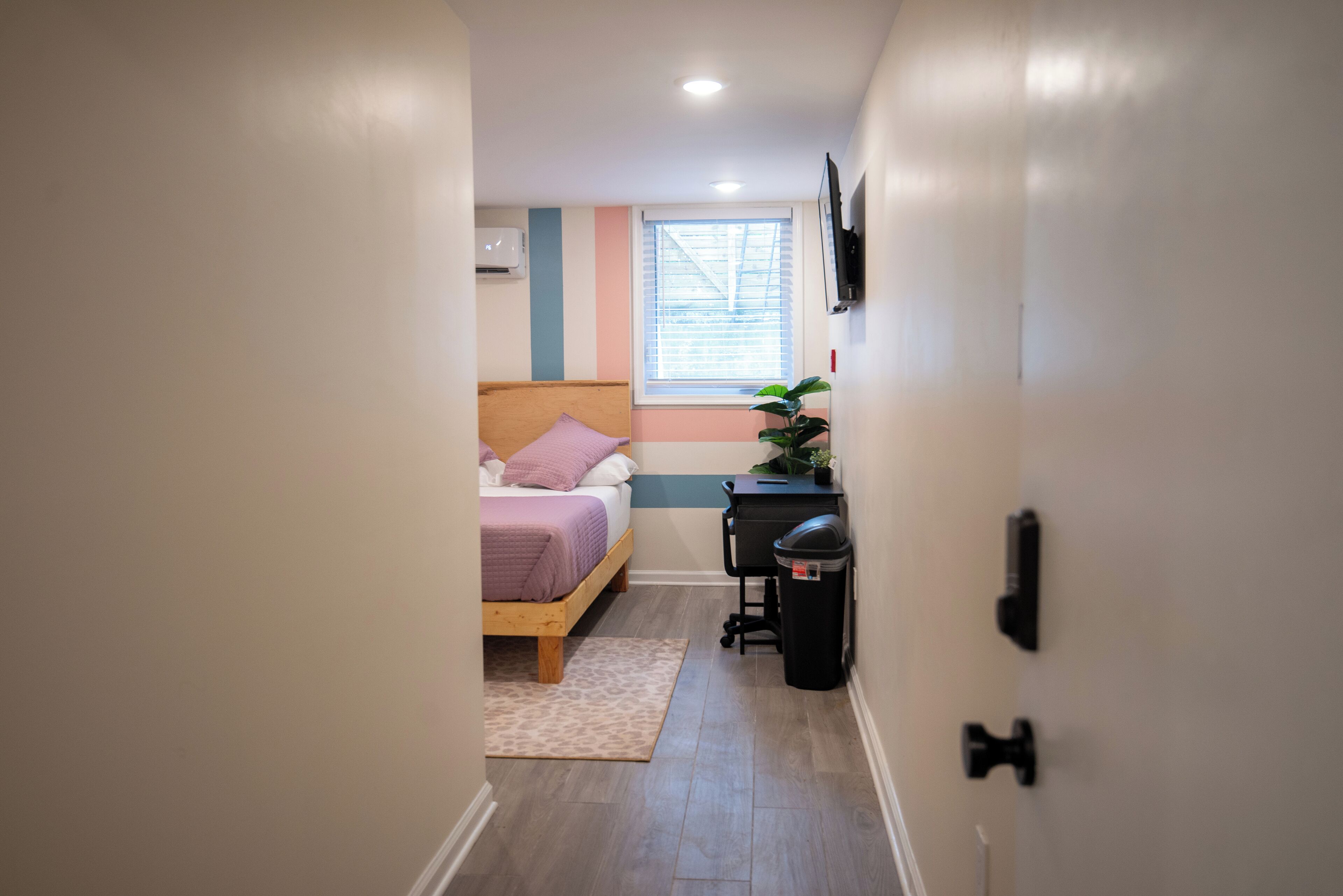 A narrow hallway leading to a bedroom with a bed, a desk, and a trash bin.