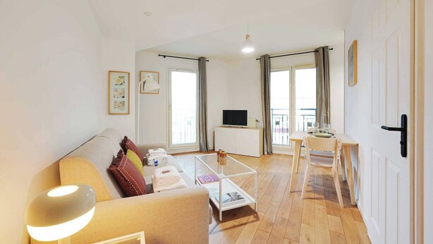 Uma sala de estar moderna com sofá, mesa de centro e TV. Há uma varanda com mesa e cadeiras.