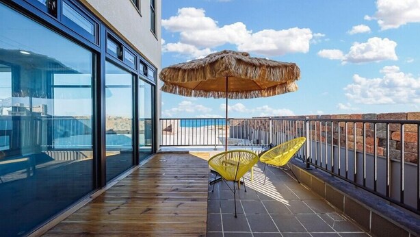 Un balcon agrémenté d’un parasol en chaume, de chaises jaunes et offrant une vue sur la plage.