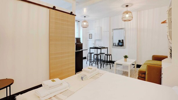 A modern hotel room with a bed, a small dining table with chairs, a desk with a chair, a sofa, and a sliding door.