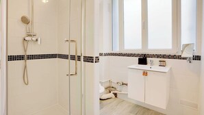 A modern bathroom with a glass-enclosed shower, a white sink with a black tiled backsplash, and a window above the sink.