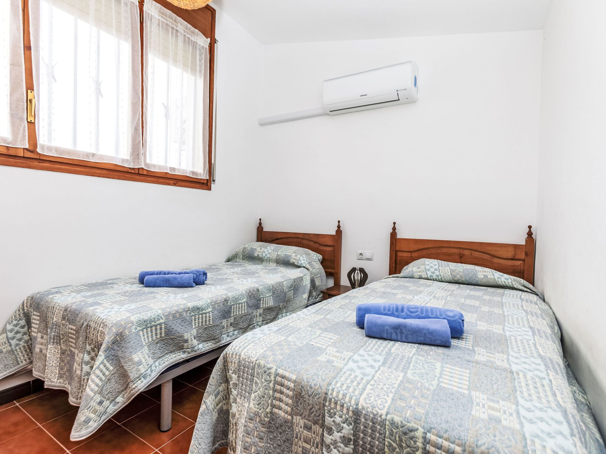 Un dormitorio con dos camas, cada una con ropa de cama estampada y toallas azules. Una unidad de aire acondicionado está montada en la pared sobre las camas.