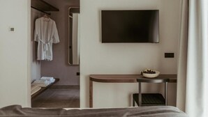 A modern hotel room with a bed, a TV mounted on the wall, a small table with a bowl of fruit, and a robe hanging on a rack. - Pure Beachfront Villa with pool (Sithonia)