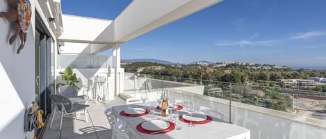 Un balcón con una mesa blanca dispuesta para comer, sillas blancas y una vista despejada del paisaje.
