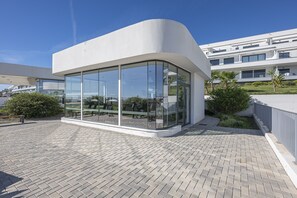 A modern building with large glass windows, a curved white facade, and a paved patio area with outdoor seating. - Luxury Penthouse with Seaview & Golf 5010 (Casares)