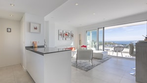 A modern kitchen with a central island, a dining area with a table and chairs, and a large sliding glass door leading to a balcony with a view of the city.