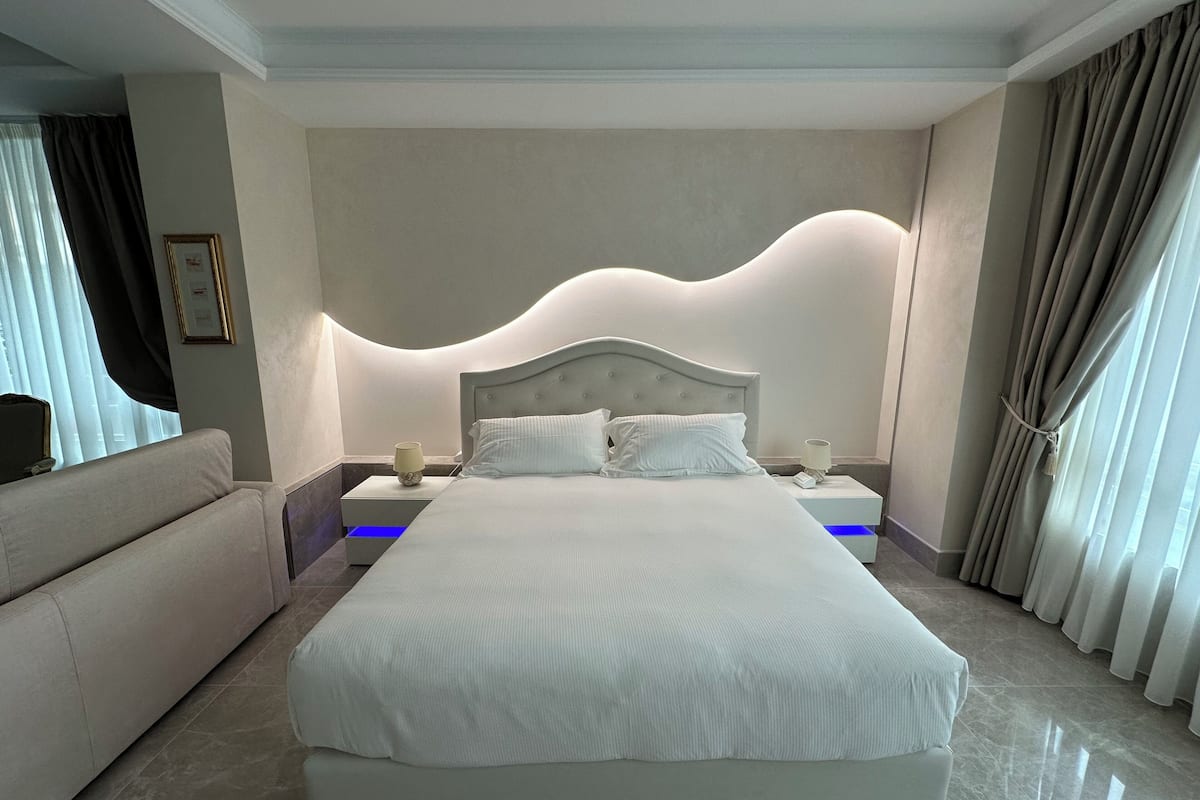A modern hotel room with a large bed, a nightstand, a lamp, and a sofa.