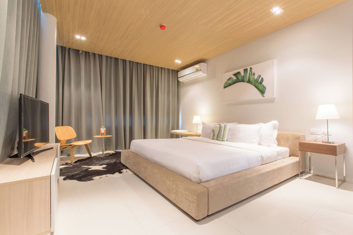 A modern hotel room with a large bed, a desk, a chair, a TV, and a small table with a lamp.