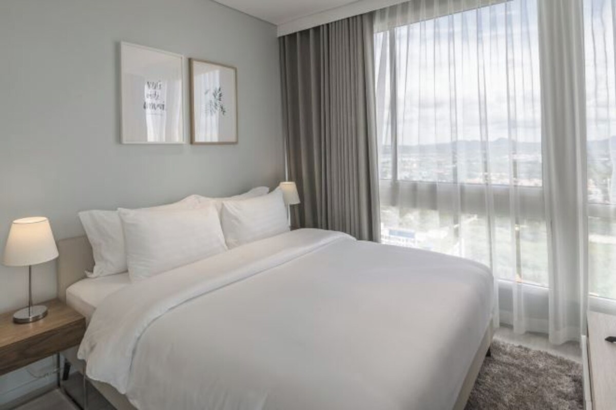 A modern hotel room with a large bed, a nightstand with a lamp, and a view of the city through sheer curtains.