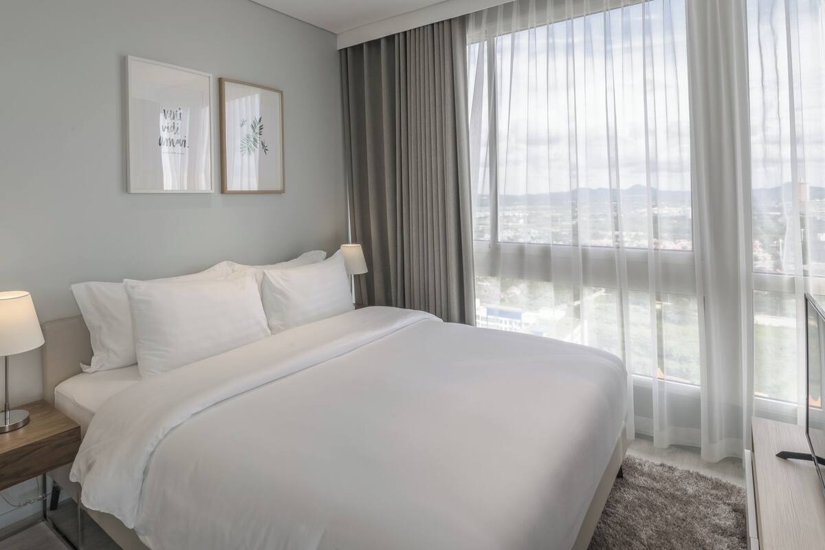 A hotel room with a large bed, a bedside table with a lamp, a view of the city through sheer curtains, and two framed pictures on the wall.