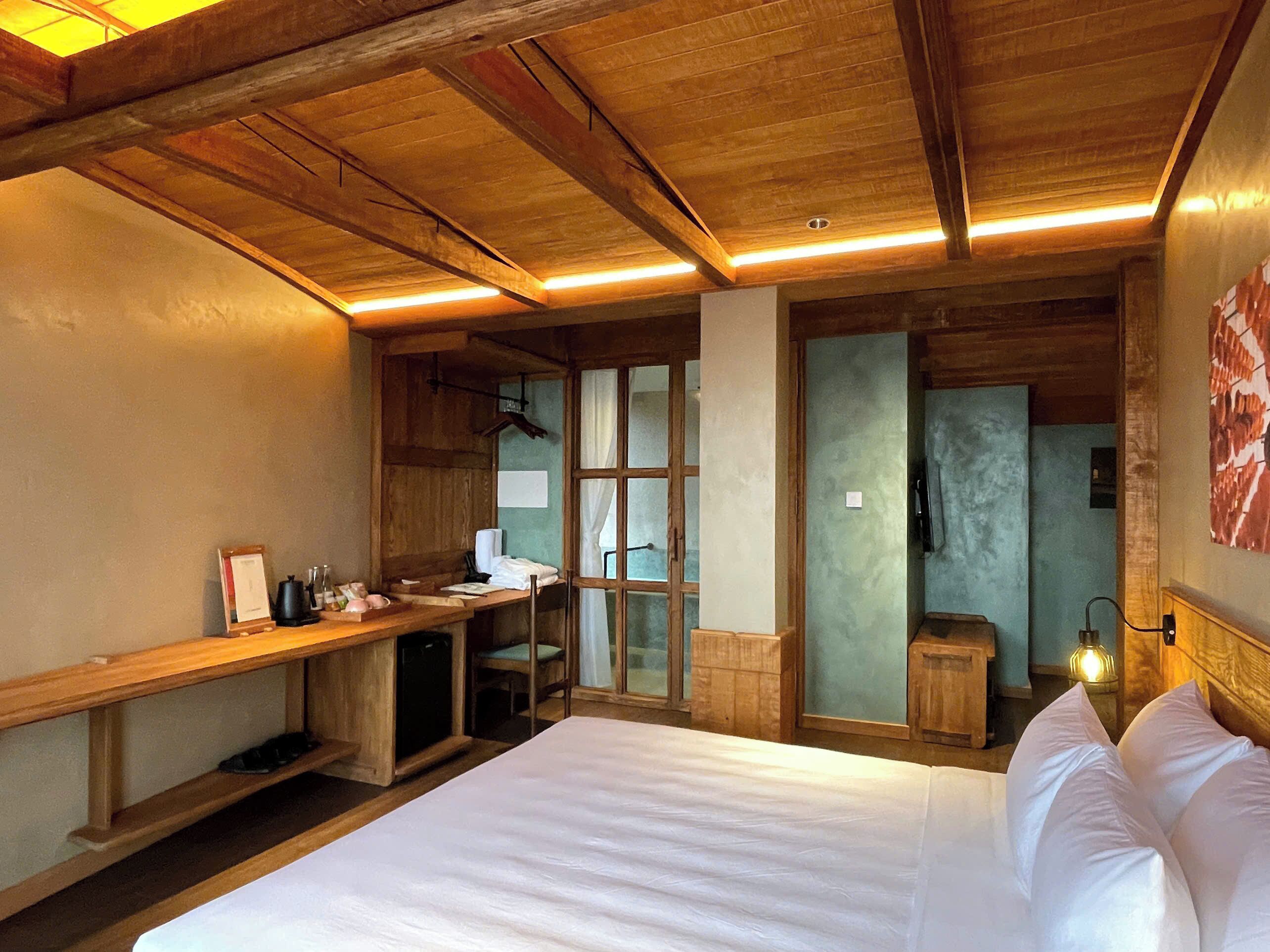 Una habitación con piso de madera, una cama, un escritorio y una silla. Se observa una ventana con cortinas, un teléfono en la pared y una lámpara.
