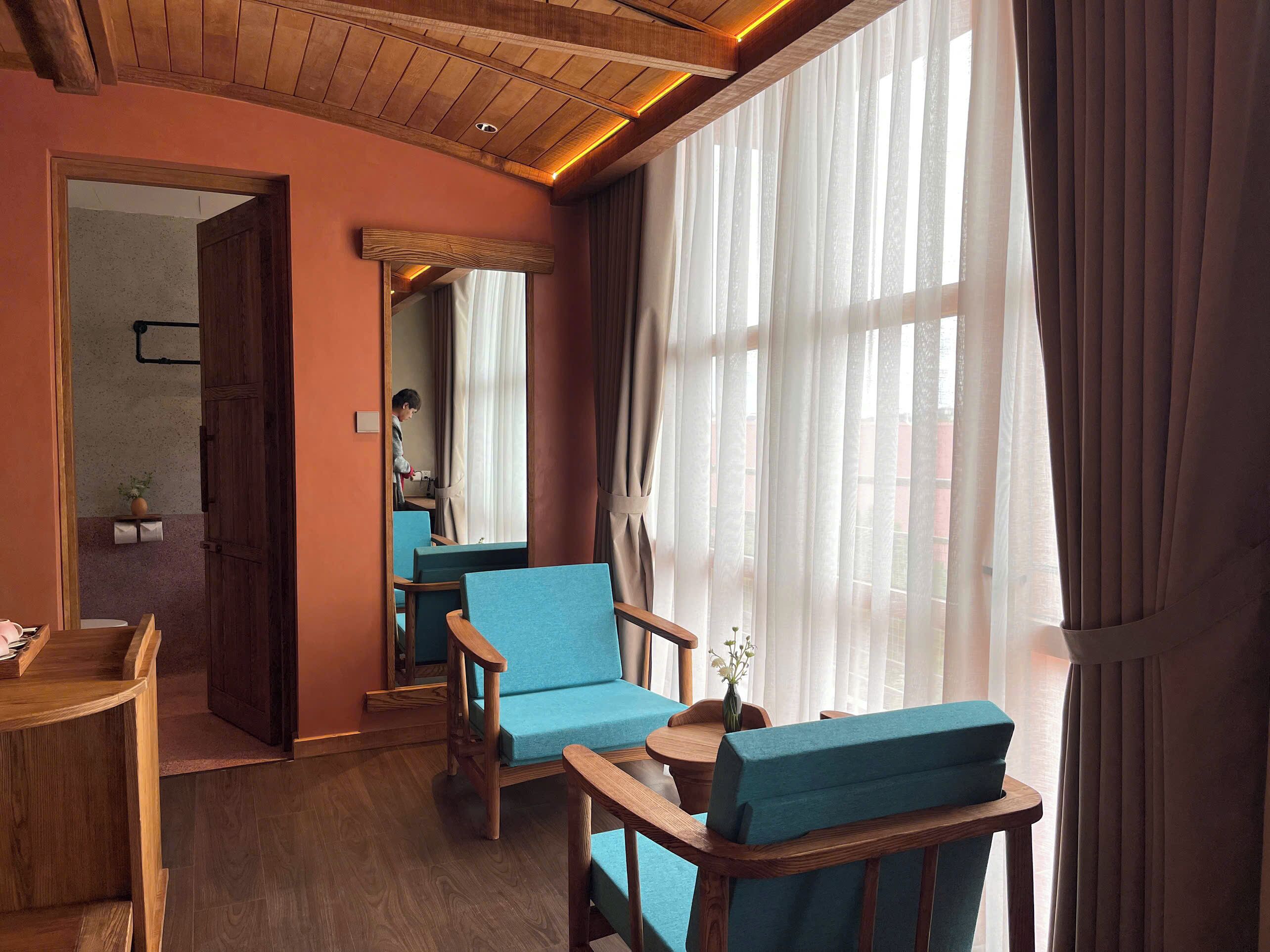 A room with wooden furniture, a large window with sheer curtains, and a visible door leading to another room.