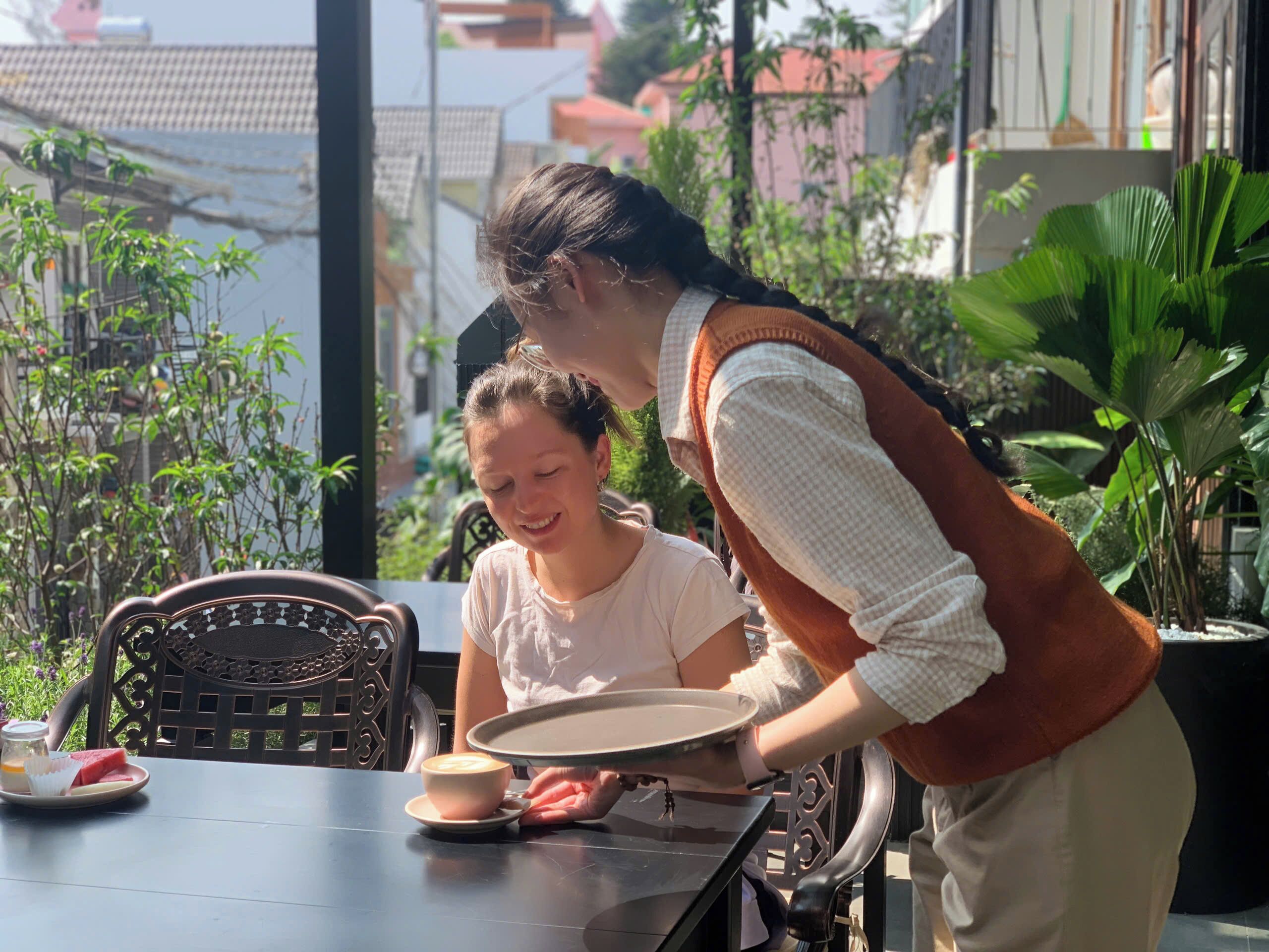 A person serving coffee and a slice of watermelon to another person at an outdoor table with a view of buildings and plants.