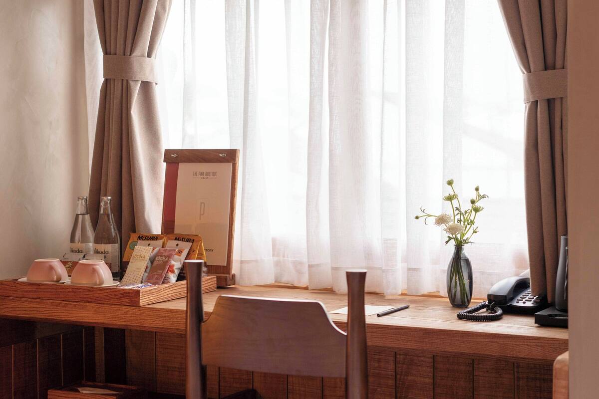 Une pièce avec un bureau en bois, une chaise, un vase de fleurs et une fenêtre avec des rideaux.