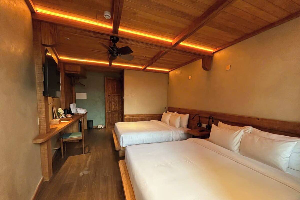 Une chambre avec deux lits, un bureau en bois, un ventilateur de plafond et un plancher en bois.