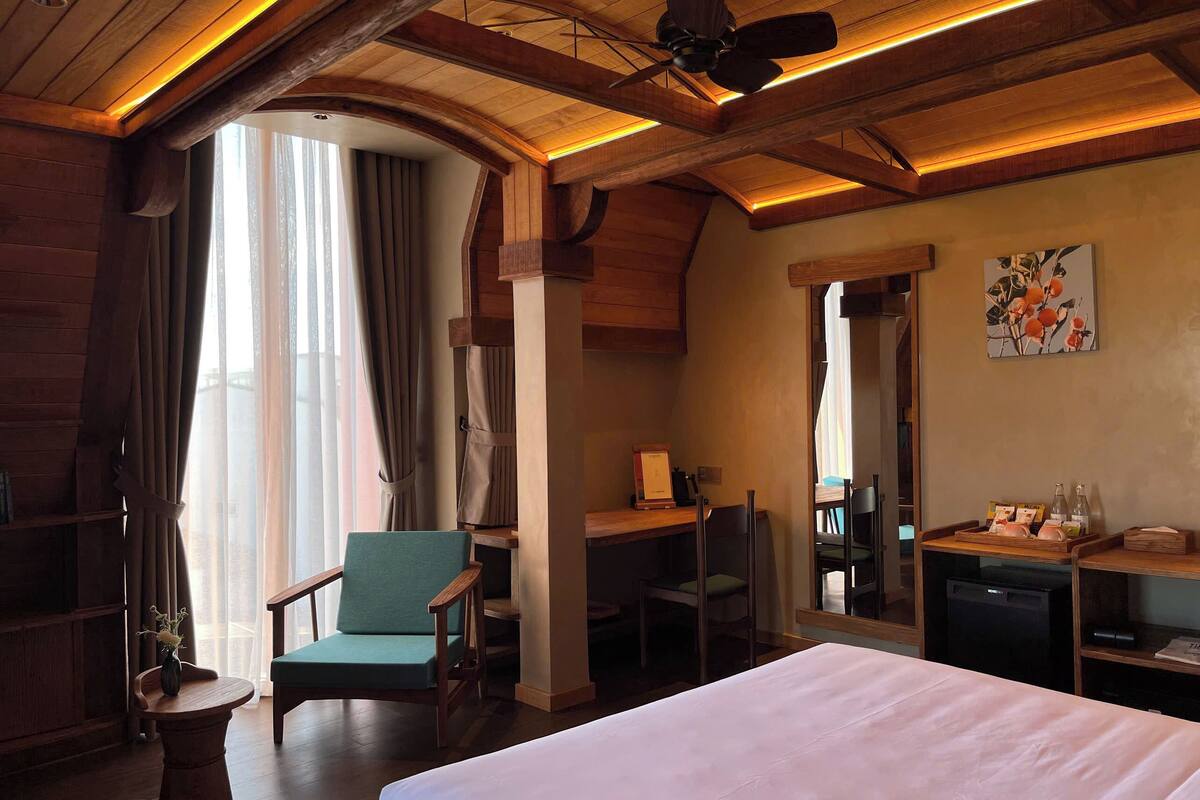Une chambre d’hôtel avec un lit, un bureau, une chaise, une petite table avec un vase et un ventilateur de plafond.