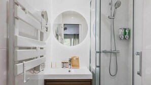 A modern bathroom with a glass shower enclosure, a round mirror, and a white vanity with toiletries. - Modern "Green Suites" residence - Meudon/Vélizy (Meudon)