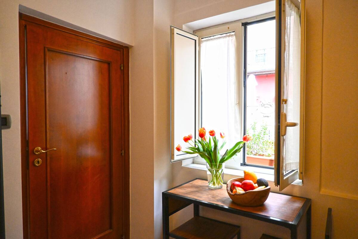 Eine Holztür, ein Fenster mit Blick auf ein Gebäude und ein kleiner Tisch mit Blumenvase und Obstschale.