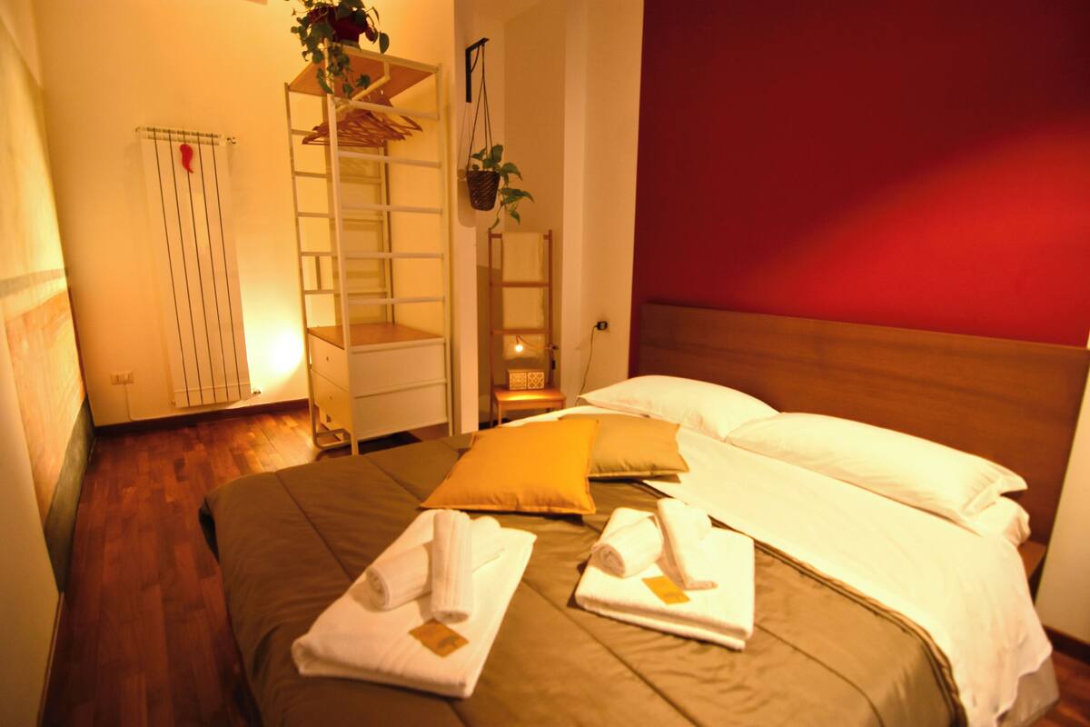 Ein Hotelzimmer mit einem Bett, zwei Kissen, einem Holz-Kopfteil, einer roten Wand, einer Pflanze, einer Leiter und einem kleinen Tisch mit einer Lampe.