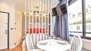 A modern hotel room with a dining area featuring a round white table, white chairs, and a small white sofa. A TV is mounted on the wall, and there is a window with a view of buildings outside. - Magnificent studio - 2P - near the Palais Garnier (Paris)