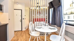 A compact room with a white table set for two, a white chair, a TV mounted on the wall, and a window with a view of buildings. - Magnificent studio - 2P - near the Palais Garnier (Paris)