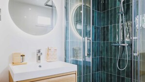 Un baño moderno con tocador blanco, espejo redondo y cabina de ducha con mampara de vidrio y ducha de mano.