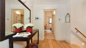 Un pasillo con suelo de madera, una consola oscura de madera con un espejo, dos jarrones rojos con flores blancas y un banco estampado.