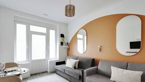 A modern living room with a grey sofa, a round mirror, and a dining area with a table set for four. - Magnificent studio - 2P - Near Bastille (Paris)