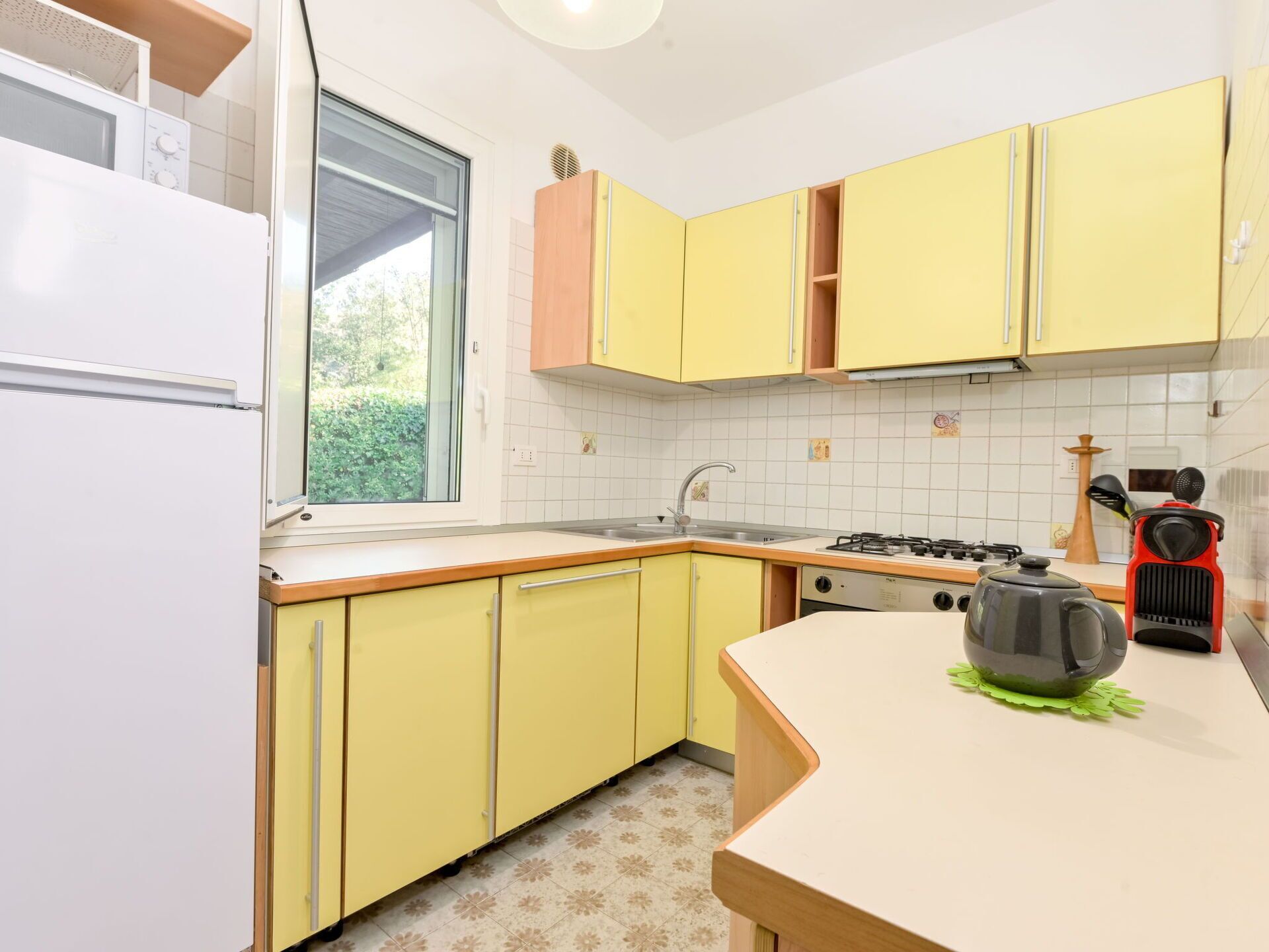 A kitchen with yellow cabinets, a white refrigerator, a window, a sink, a stove, and a kettle on the counter.
