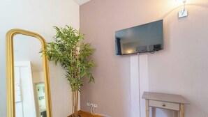 Una habitación con un espejo grande, una planta en maceta, un televisor de pantalla plana montado en la pared y una pequeña mesa de madera con cajón.