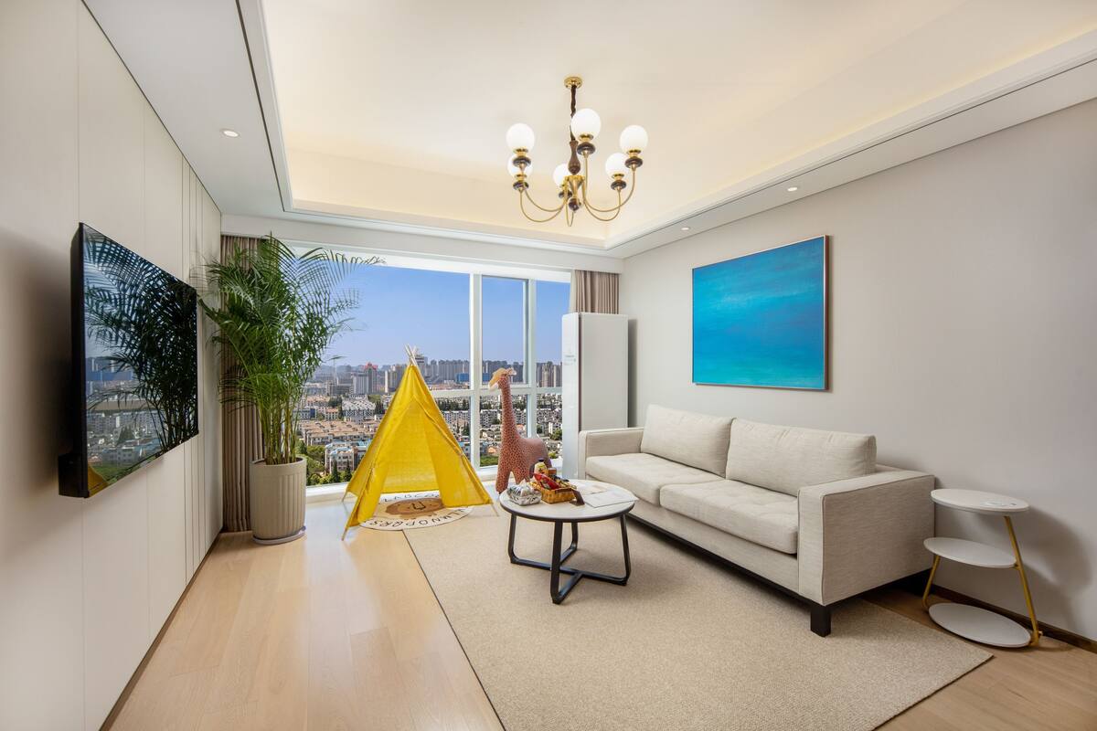 Ein modernes Wohnzimmer mit einer Couch, einem kleinen Couchtisch, einem gelben Spielzelt und Blick auf die Stadt durch große Fenster.