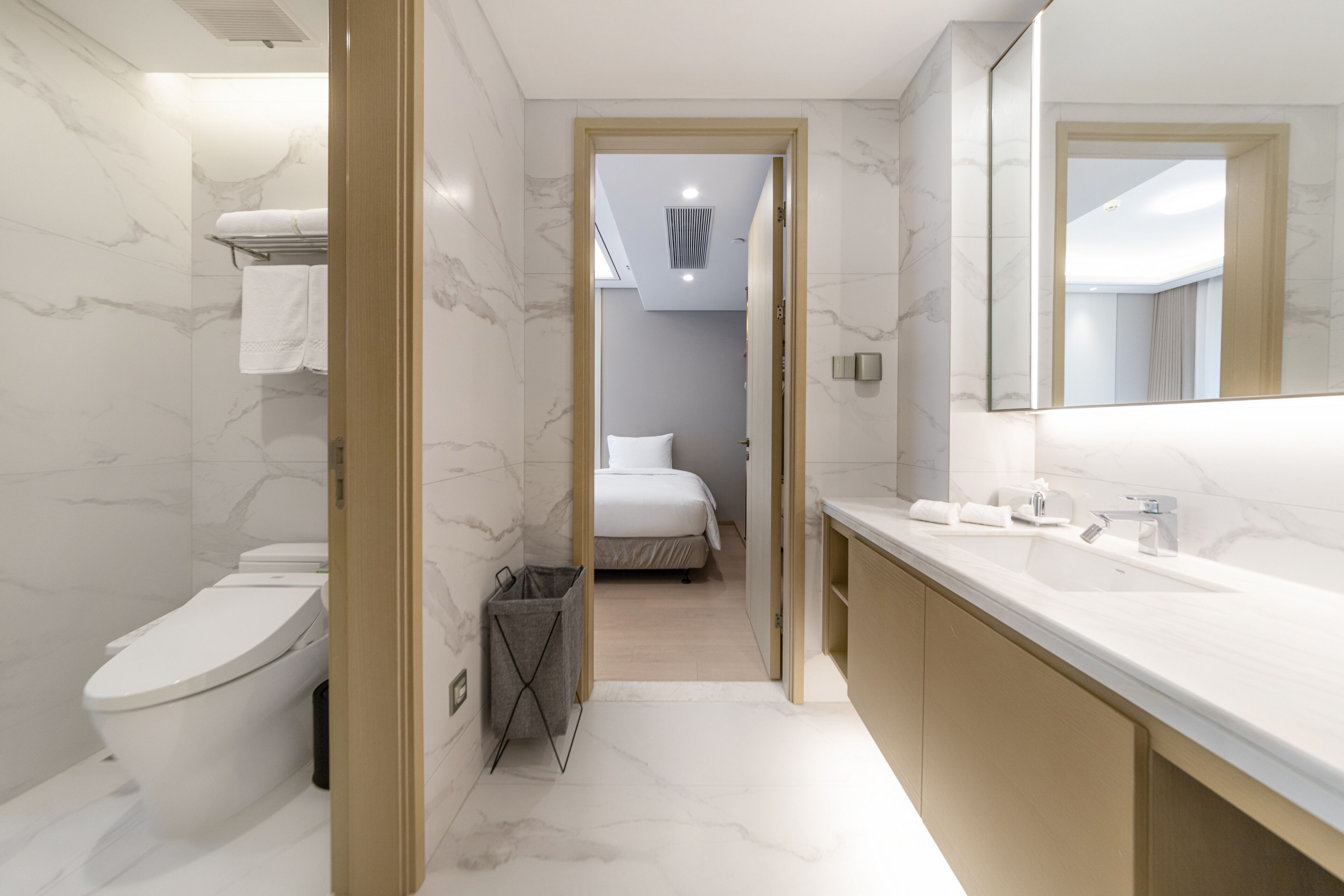 Une salle de bain moderne avec des toilettes, un lavabo et un miroir. Un couloir mène à une chambre à coucher équipée d’un lit et d’un luminaire au plafond.