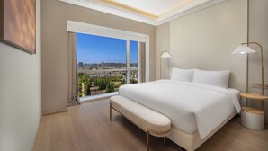 Une chambre d’hôtel moderne dotée d’un grand lit, d’une banquette, de deux tables de chevet et offrant une vue sur le paysage urbain grâce à de grandes fenêtres.