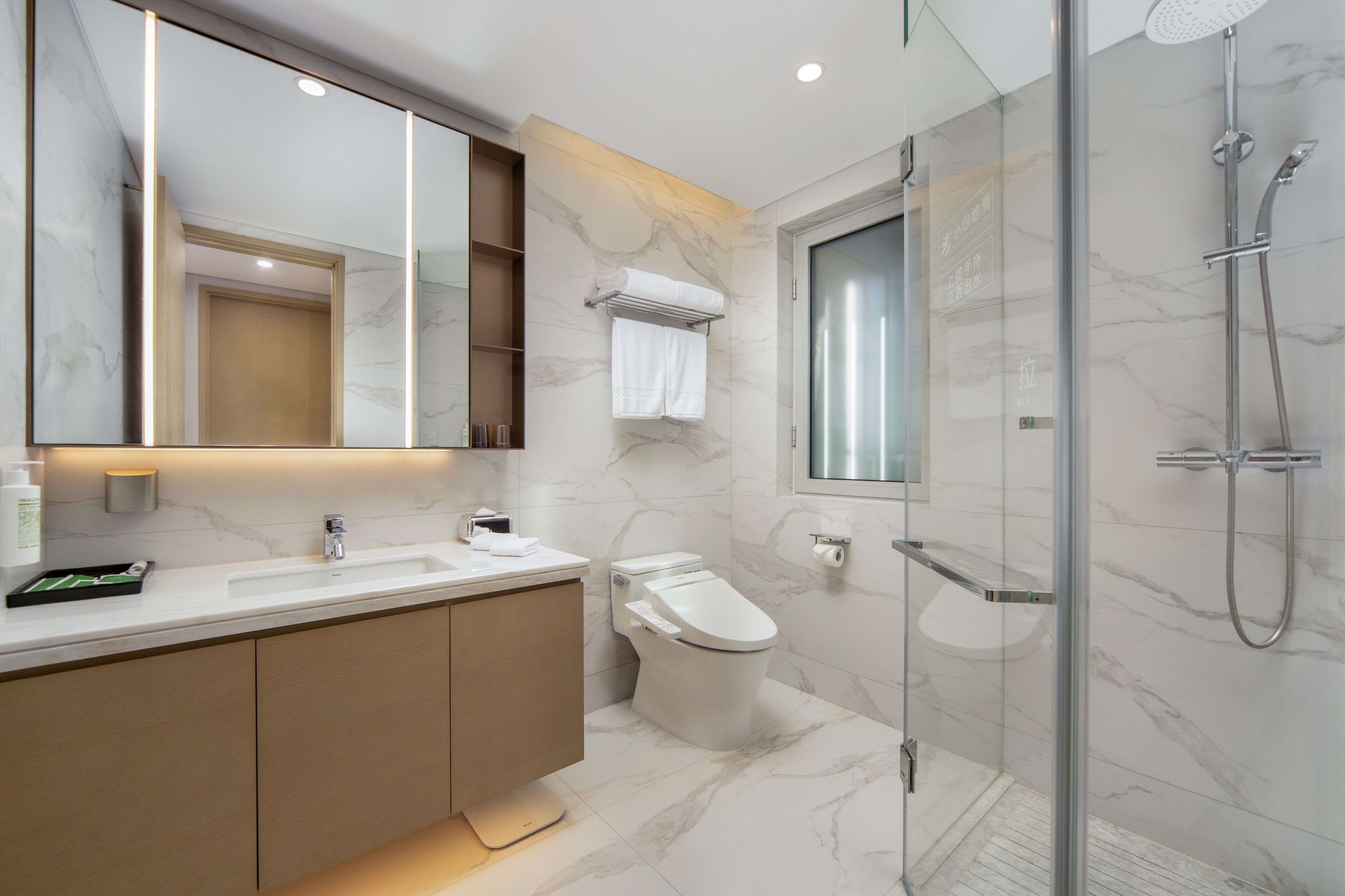 Un baño moderno con ducha envuelta en cristal, piso de mármol y un espejo grande con iluminación integrada.