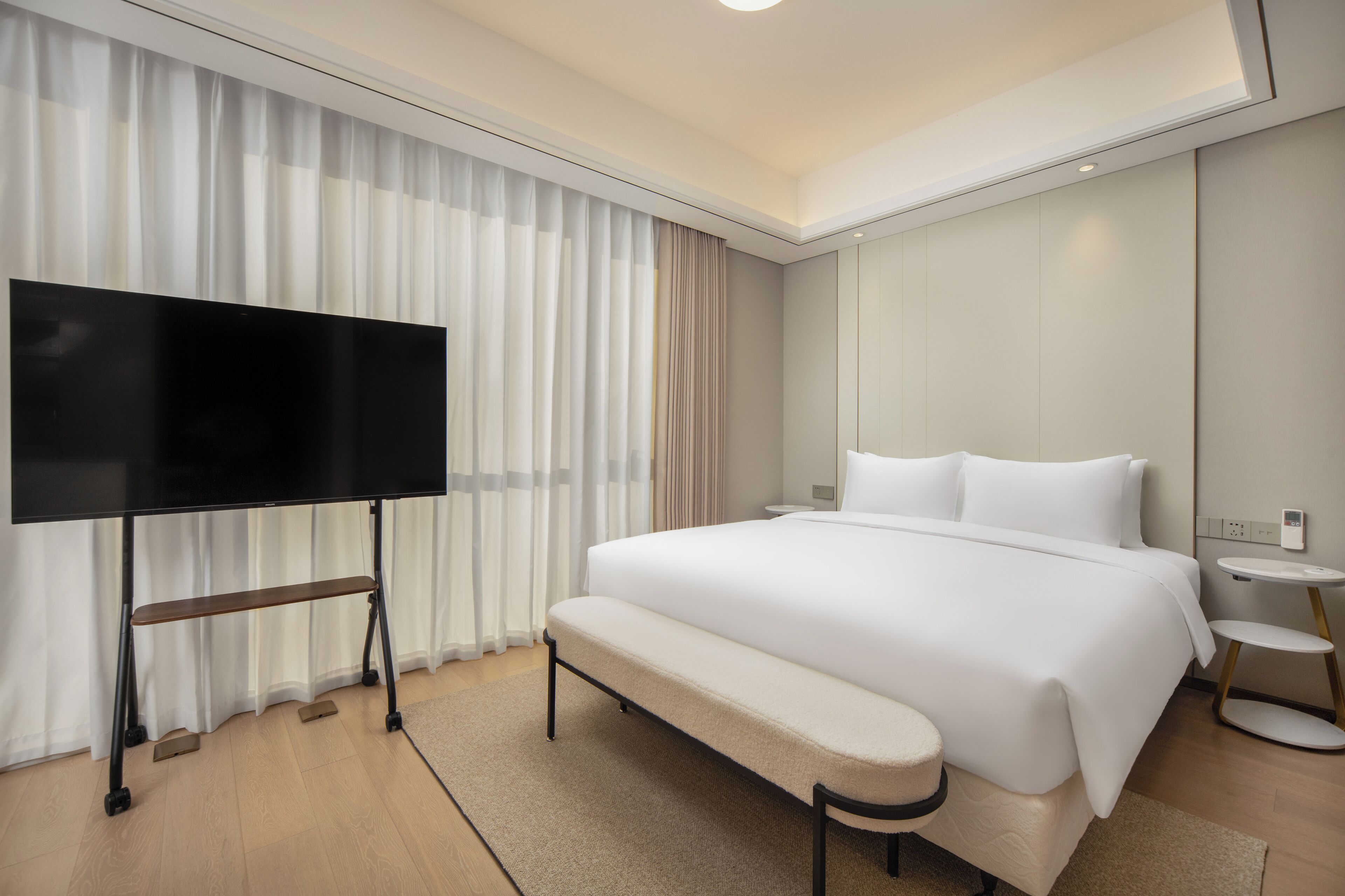 A modern hotel room with a large bed, a bench, a TV on a stand, and a bedside table.