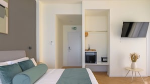 Et hotelværelse med en seng, et fjernsyn ophængt på væggen, et lille bord med en vase blomster og en dør til et badeværelse.