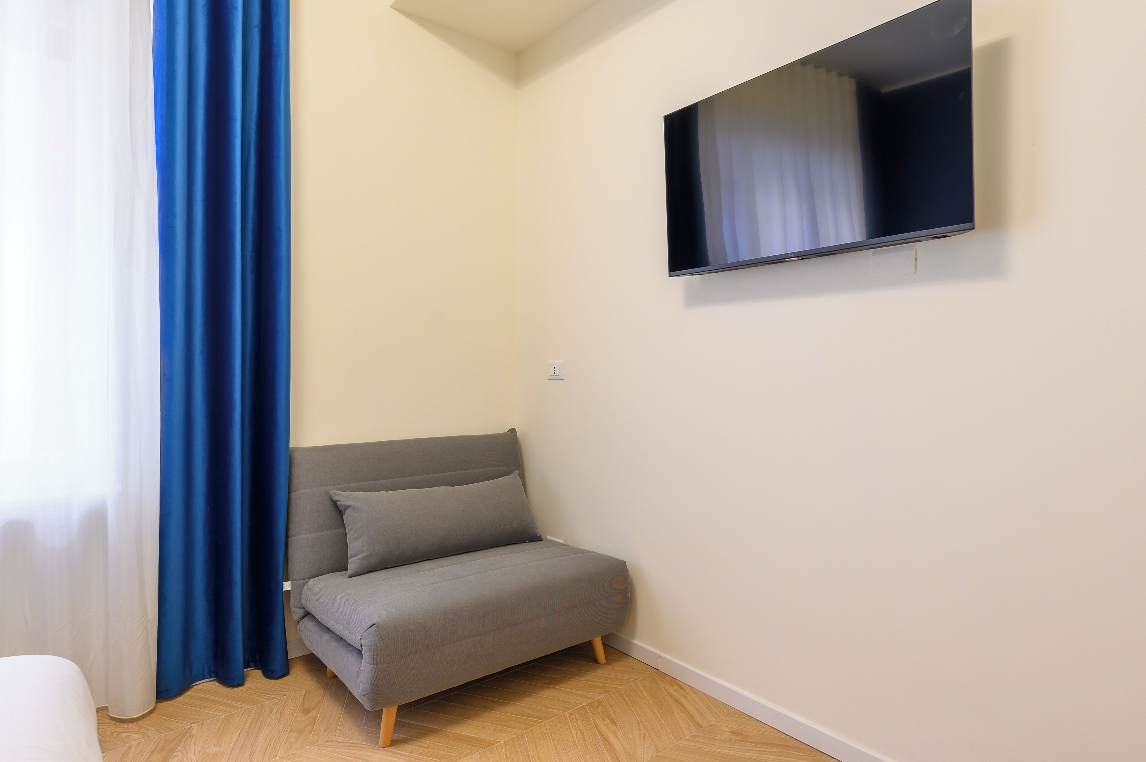 A small, modern room with a grey sofa, a flat-screen TV mounted on the wall, and a window with blue curtains.